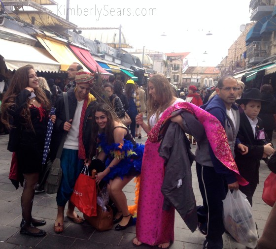 Israeli youth sport their none-too-conservative Purim costumes while a man and an Orthodox boy hurry past on a street in Mahane Yehuda.
